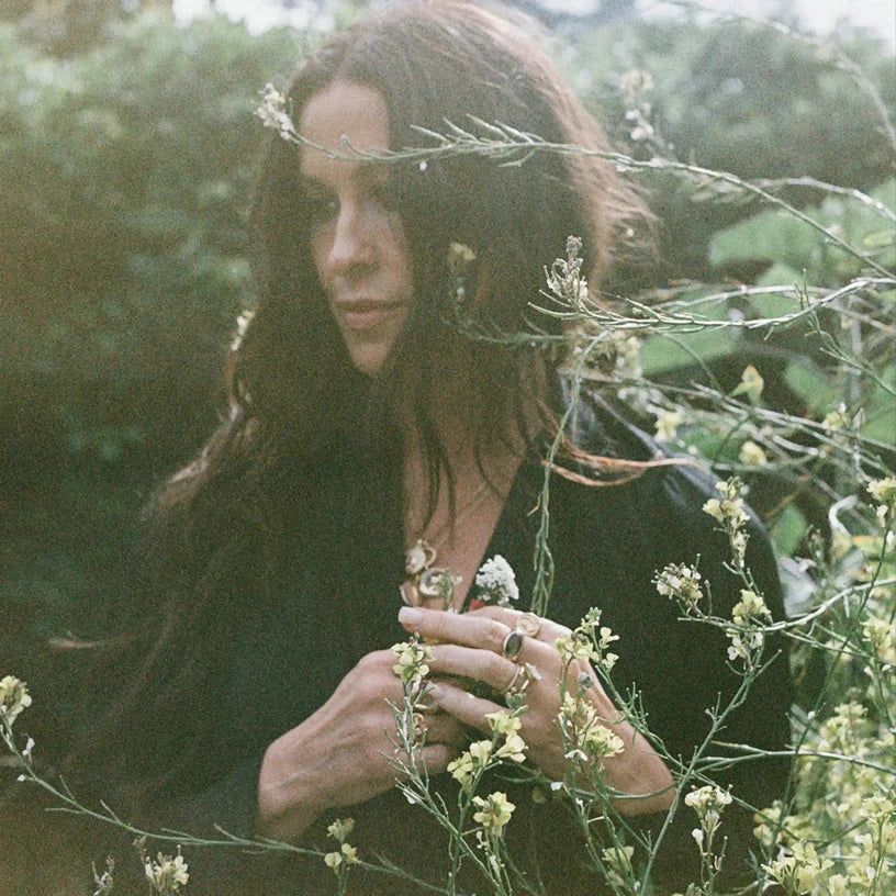A woman with long dark hair, wearing a dark top and ornate jewelry, surrounded by light green foliage and small white flowers.